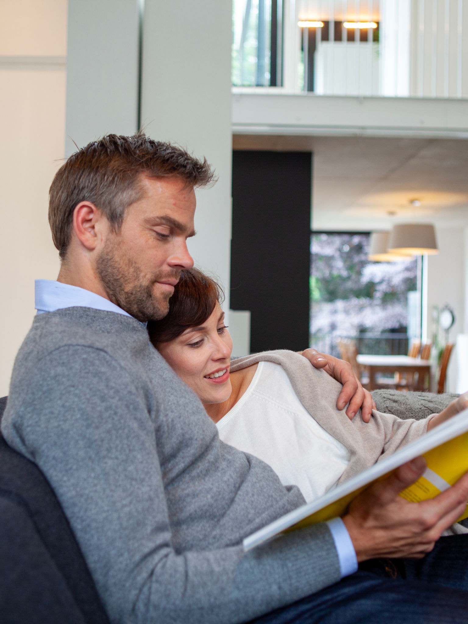 Cuplu râzând și relaxându-se pe canapea în timp ce citesc o carte la temperatura perfectă