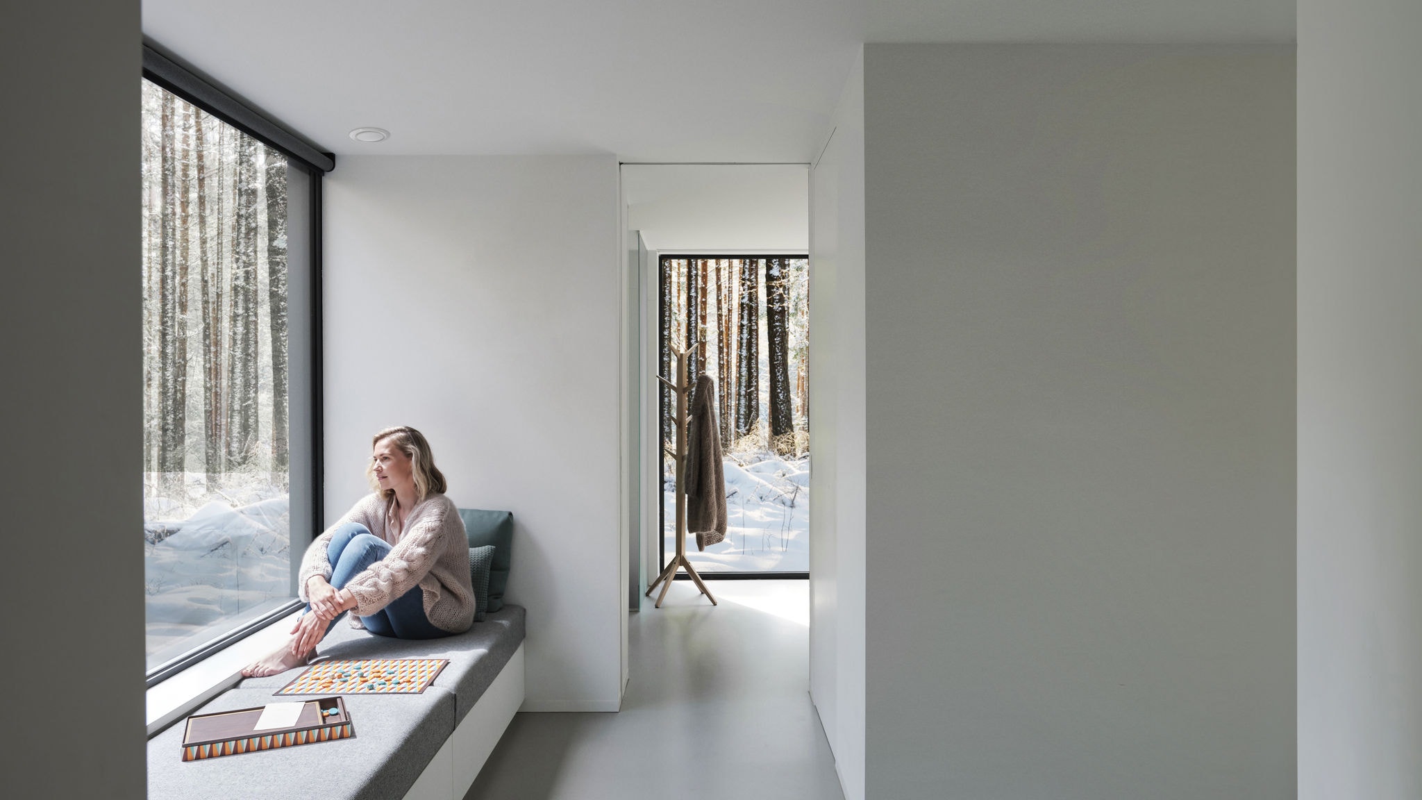 Cadru larg al unei femei care stă desculță în fața unei ferestre, privind spre copaci și zăpadă.