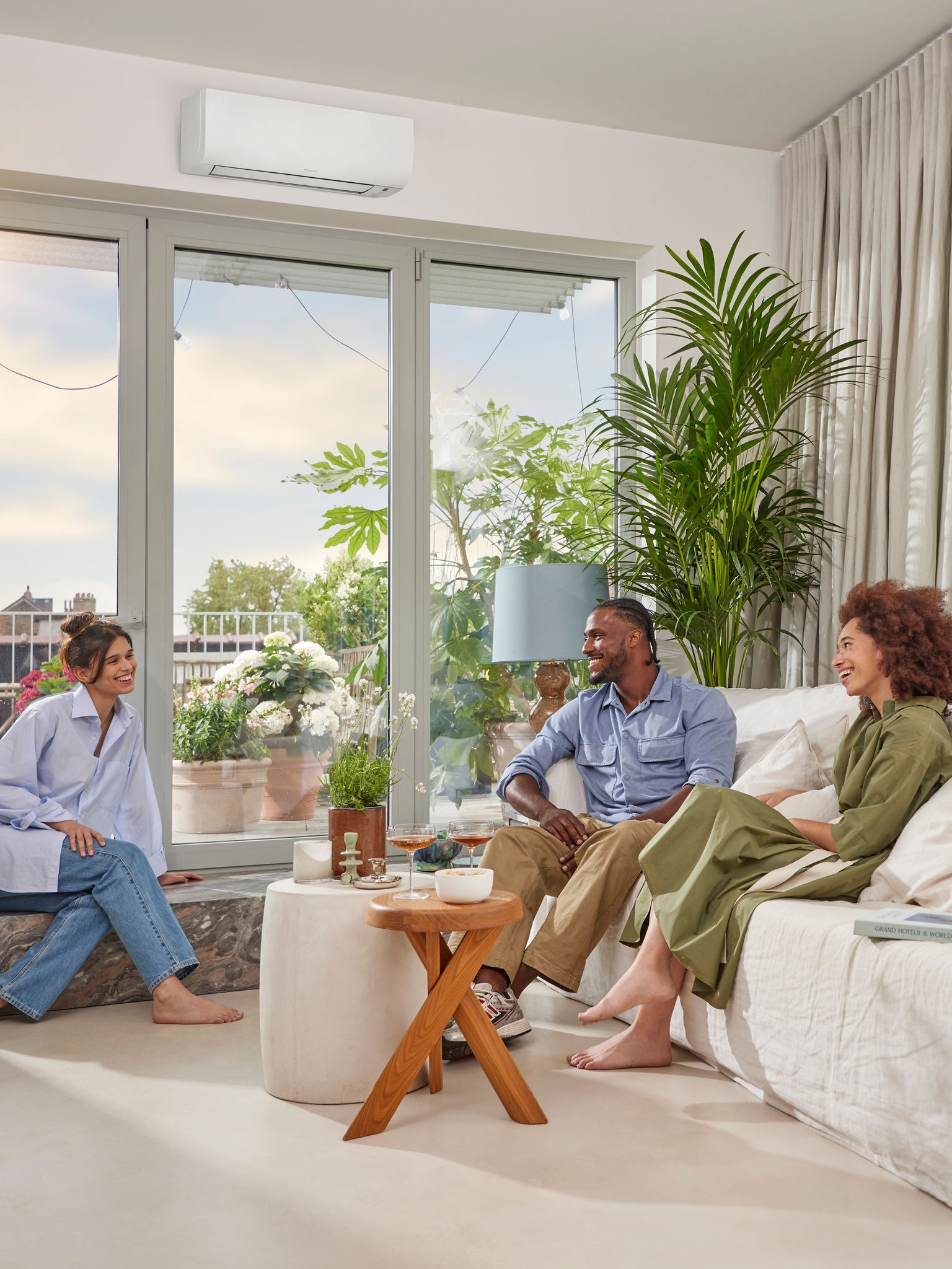 Un bărbat și două femei stau de vorbă într-o cameră luminoasă și aerisită, cu pereți albi și o pompă de căldură albă, discretă, montată pe perete, deasupra unei ferestre mari panoramice, într-o zi însorită