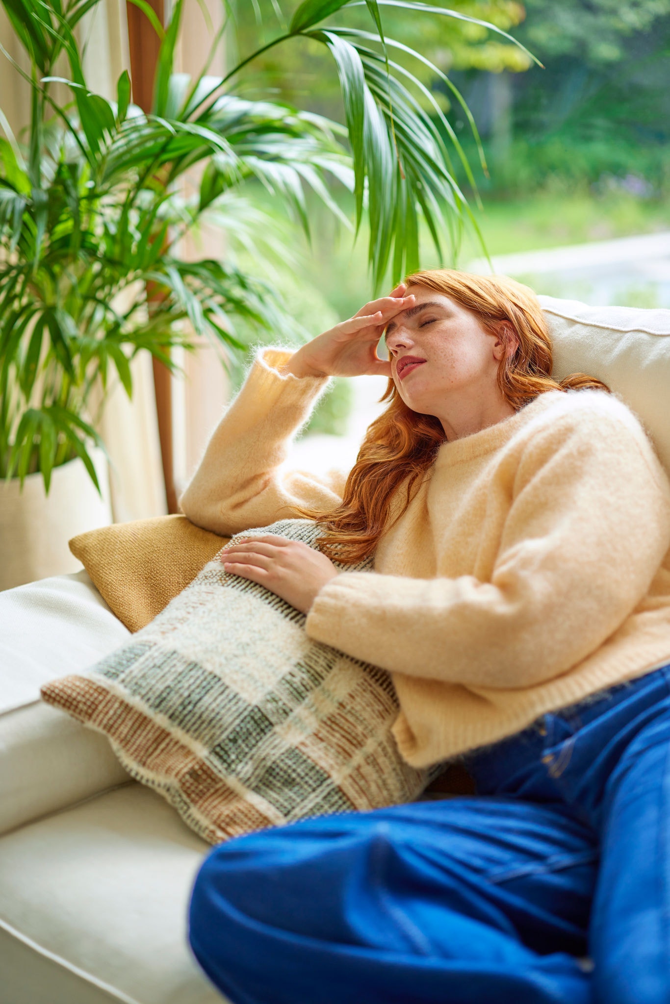 Tânără care nu se simte bine, stând pe canapea cu durere de cap, arătând impactul calității proaste a aerului și necesitatea unei ventilații corespunzătoare