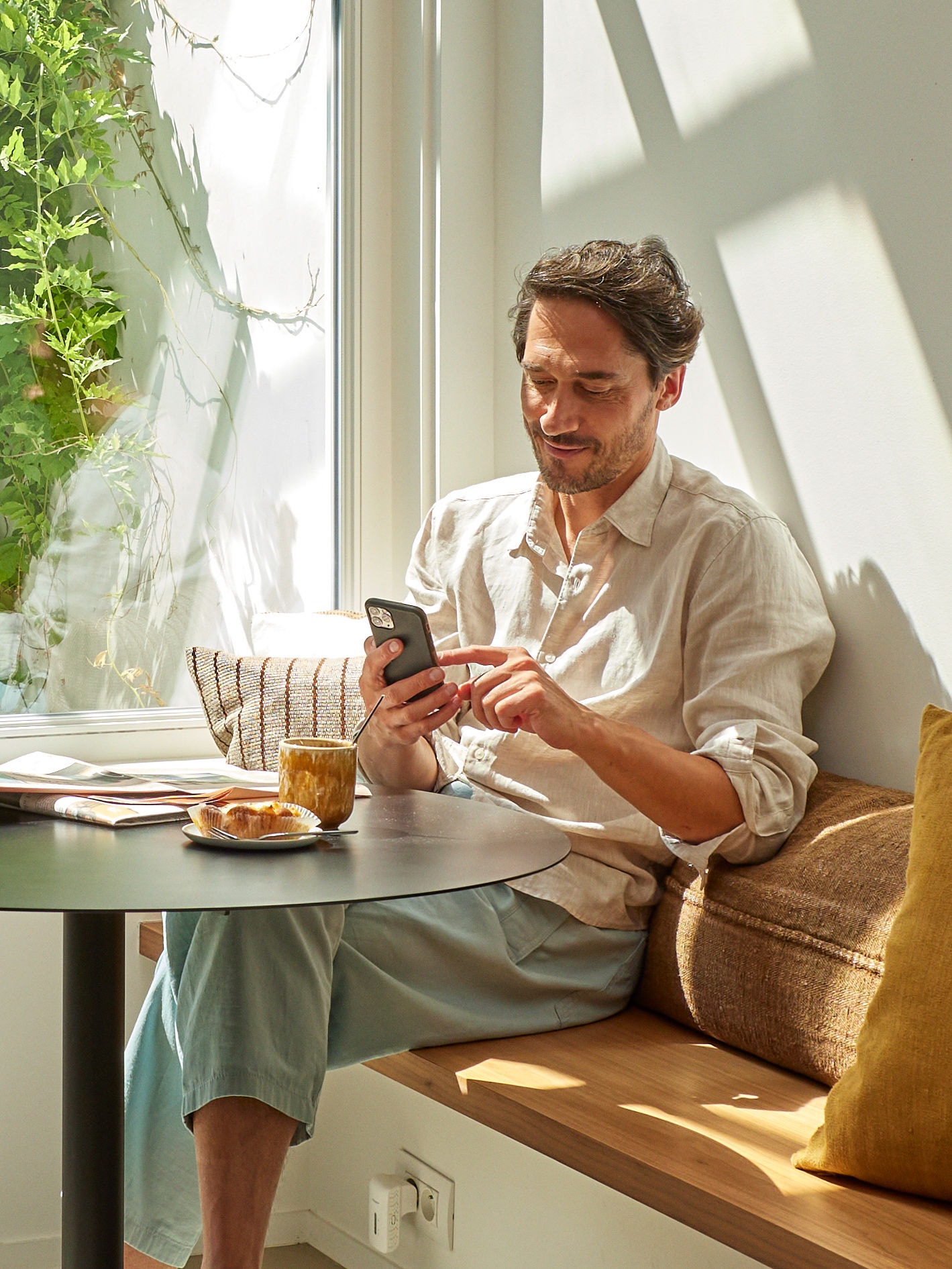 Bărbat relaxându-se și privind telefonul, așezat la o masă cu o ceașcă de cafea și mâncare în față