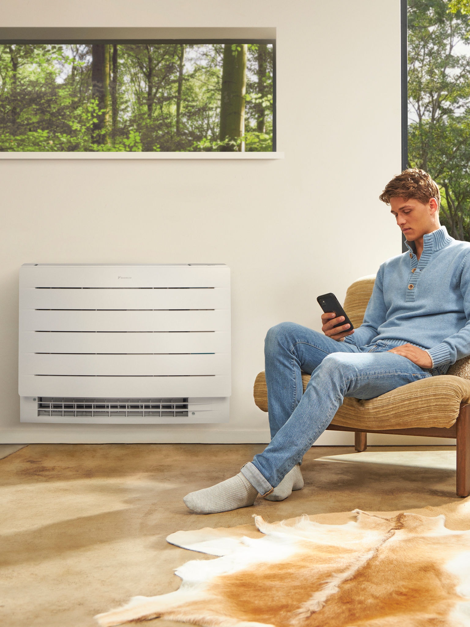 Bărbat așezat într-o cameră luminoasă, uitându-se la telefon, cu o pompă de căldură montată pe peretele din spatele lui