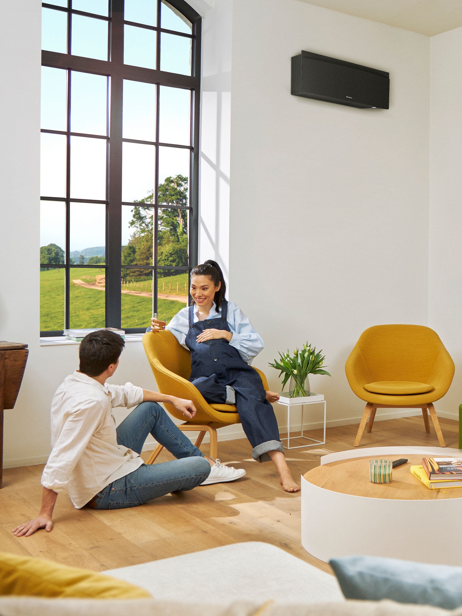 Un bărbat și o femeie stând în fața unei ferestre mari, într-o cameră luminoasă, cu o pompă de căldură neagră, montată pe peretele din spatele lor