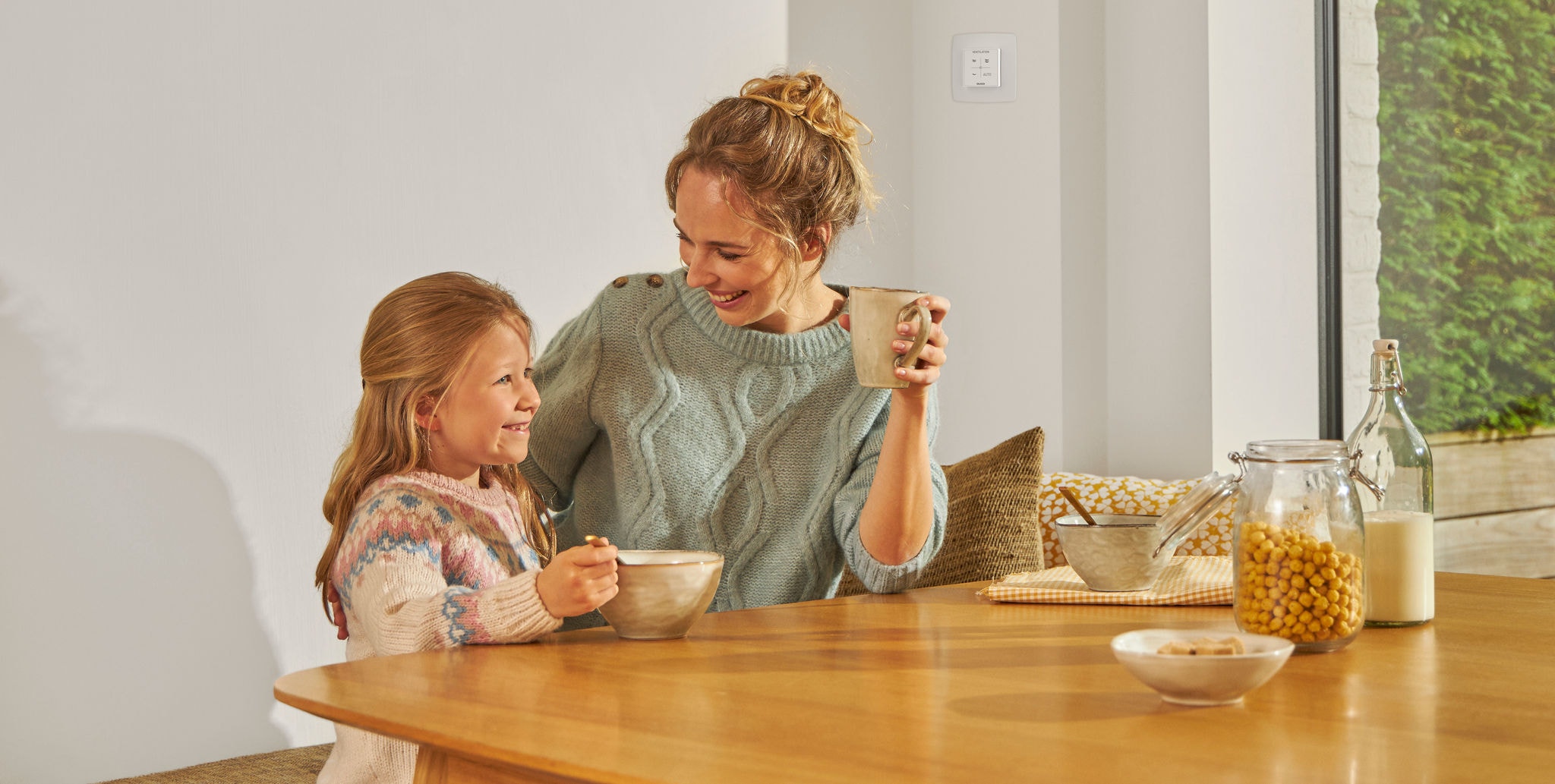 Fată tânără mâncând dintr-un bol, așezată la o masă lângă o femeie cu o cană în mână, ambele zâmbind, fericite și relaxate, bucurându-se de aer sănătos.