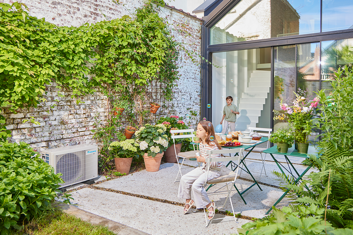 Copii care se joacă cu bile pe terasa grădinii, lângă unitatea exterioară a unei pompe de căldură.