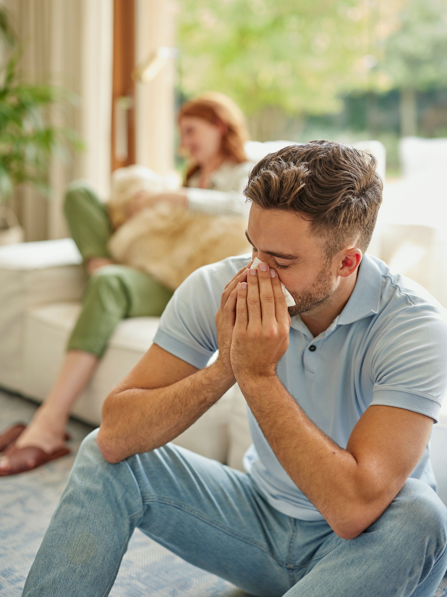 Femeie așezată pe mobilier tapițat cu un golden retriever în poale, bărbat în prim-plan strănutând