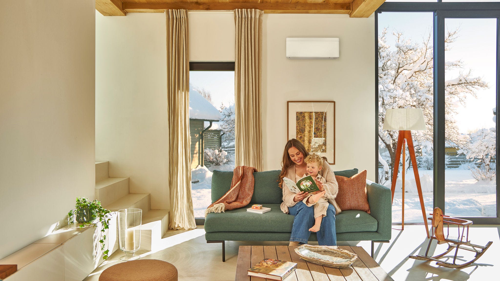 Mamă stând pe canapea sub o pompă de căldură aer-aer de perete citind o carte cu povești pentru copii și ținând copilașul în brațe, bucurându-se de căldură în timp ce afară ninge